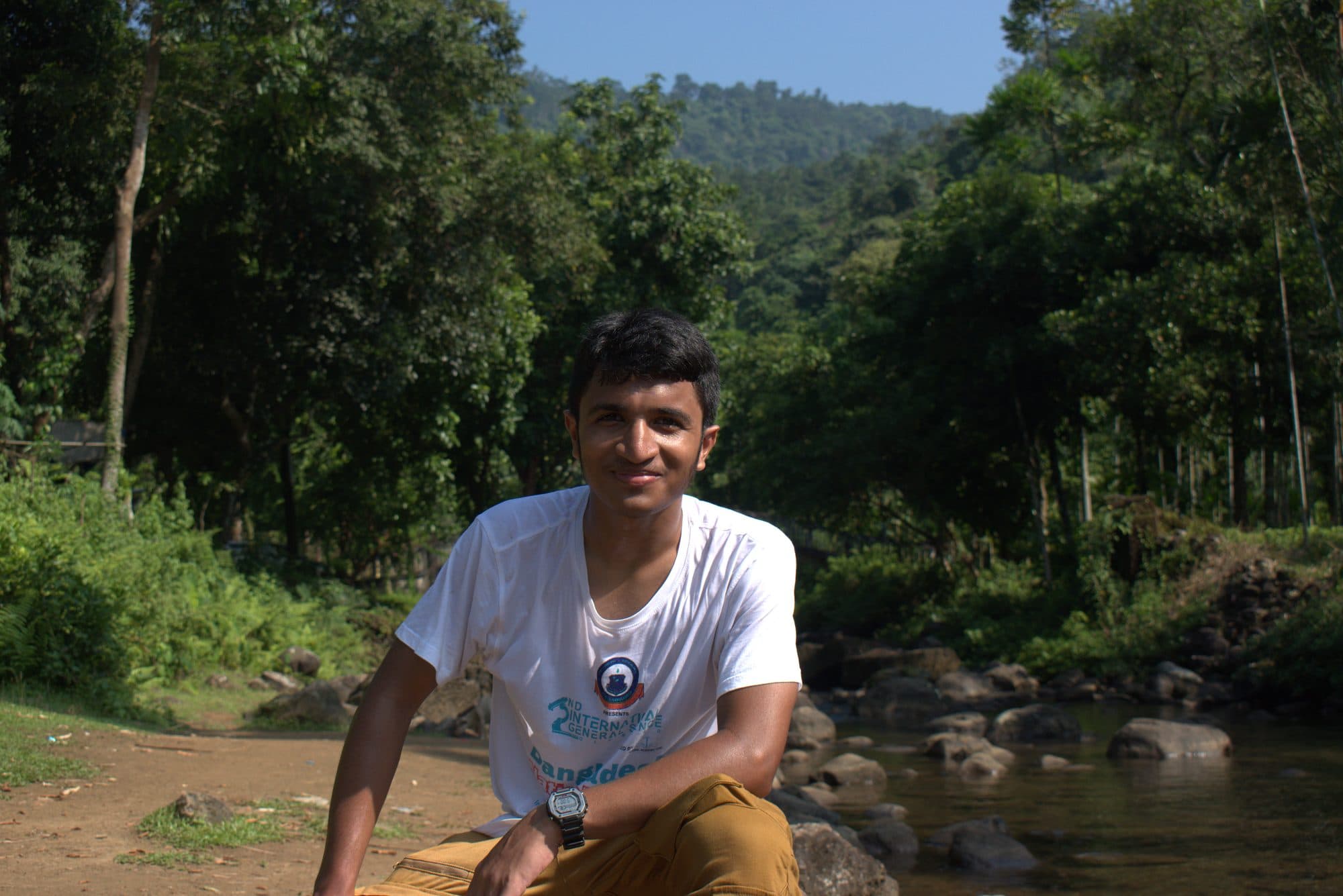 Mohammad Nasir sitting on rocks by a forest river in Sylhet